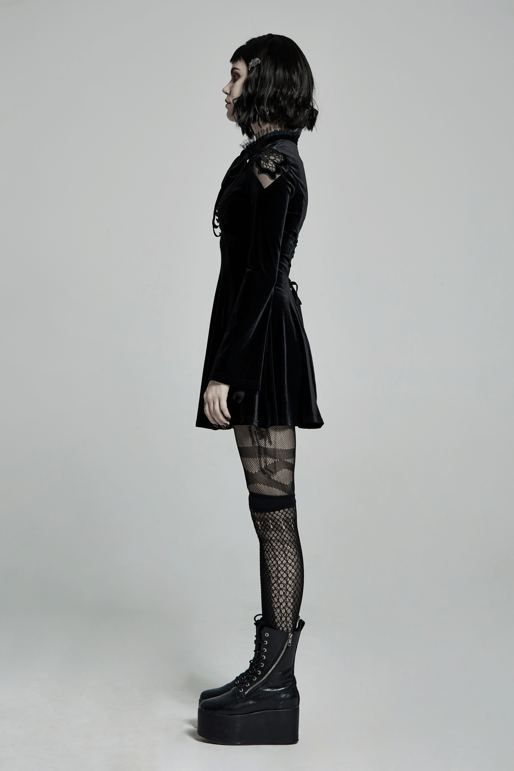 Side view of a woman in a gothic velvet mini dress with lace collar and combat boots, showcasing punk elegance.