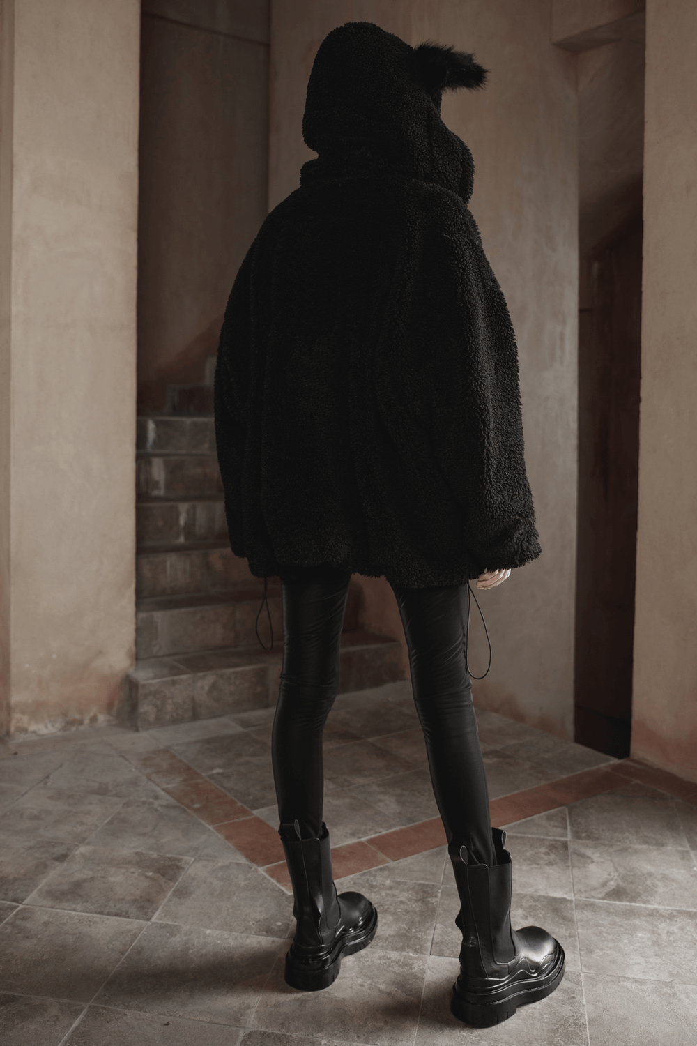 Back view of a woman in a black fluffy hooded fox ear jacket, paired with sleek leggings and stylish boots.