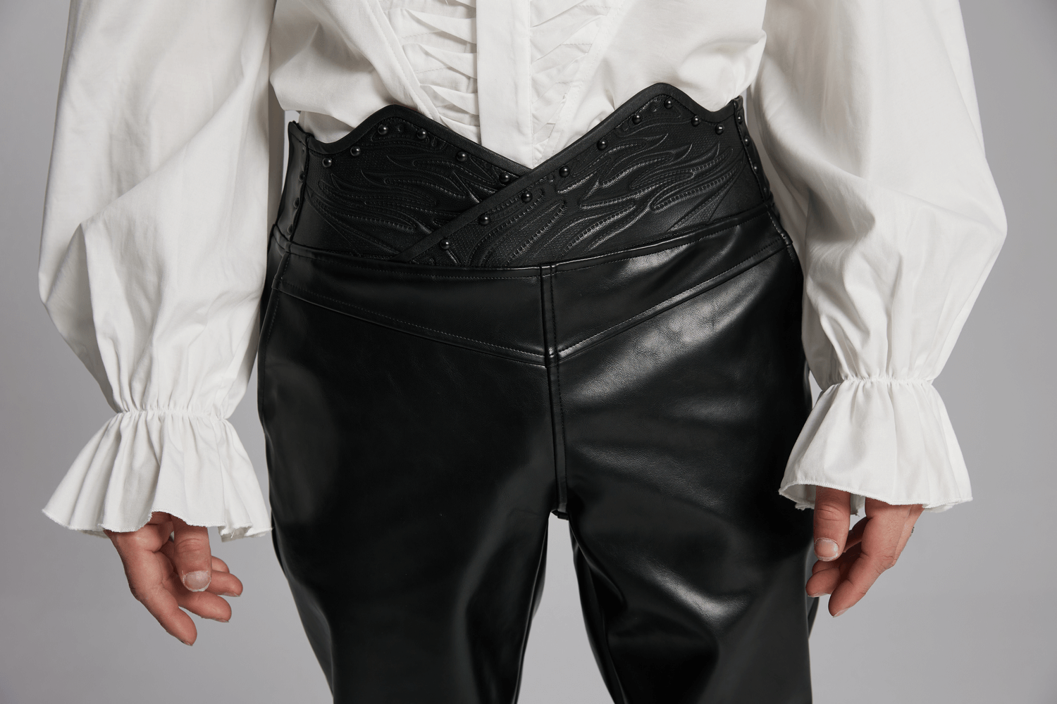 Male model showcasing black faux leather pants with a unique 3D curved waistband and studded details, styled with a white shirt.