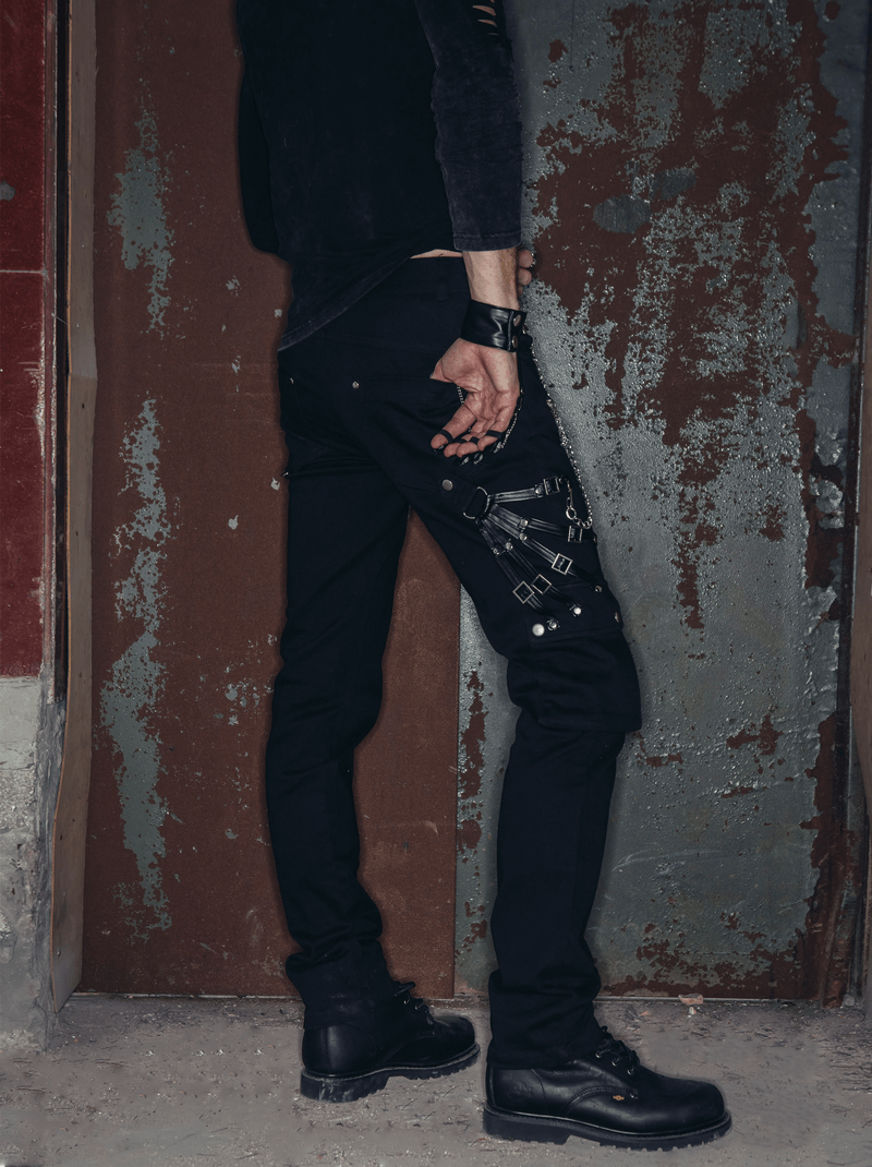 Stylish man wearing black punk rock straight long pants with chains and zippers, set against a rustic background.