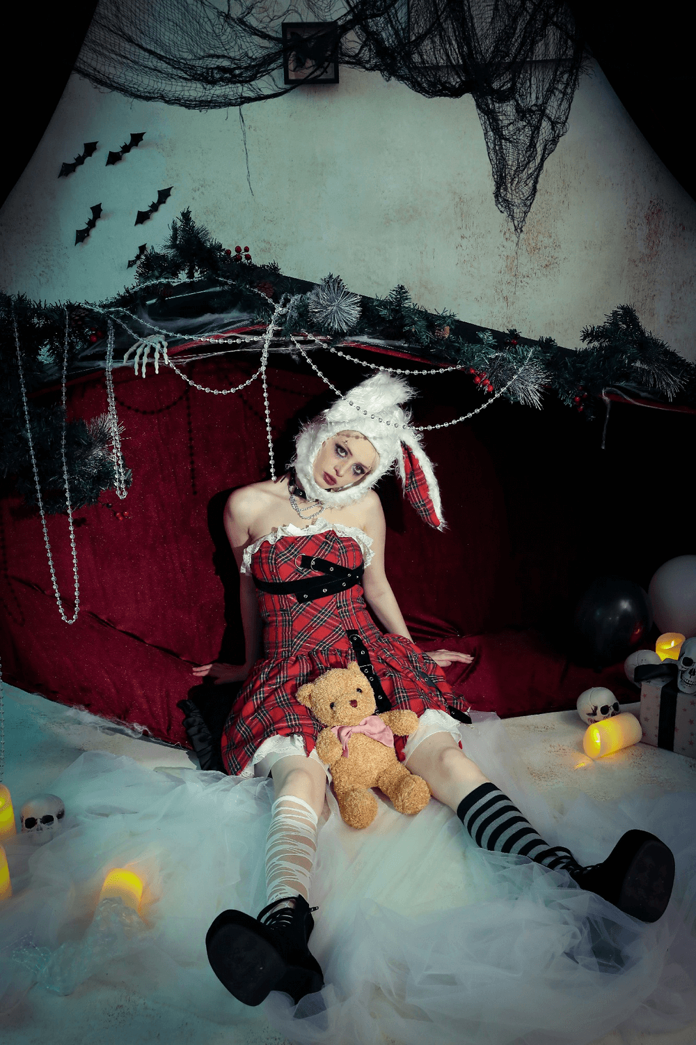 model in plaid corset dress with punk straps, sitting with teddy bear in a spooky, decorated setting.