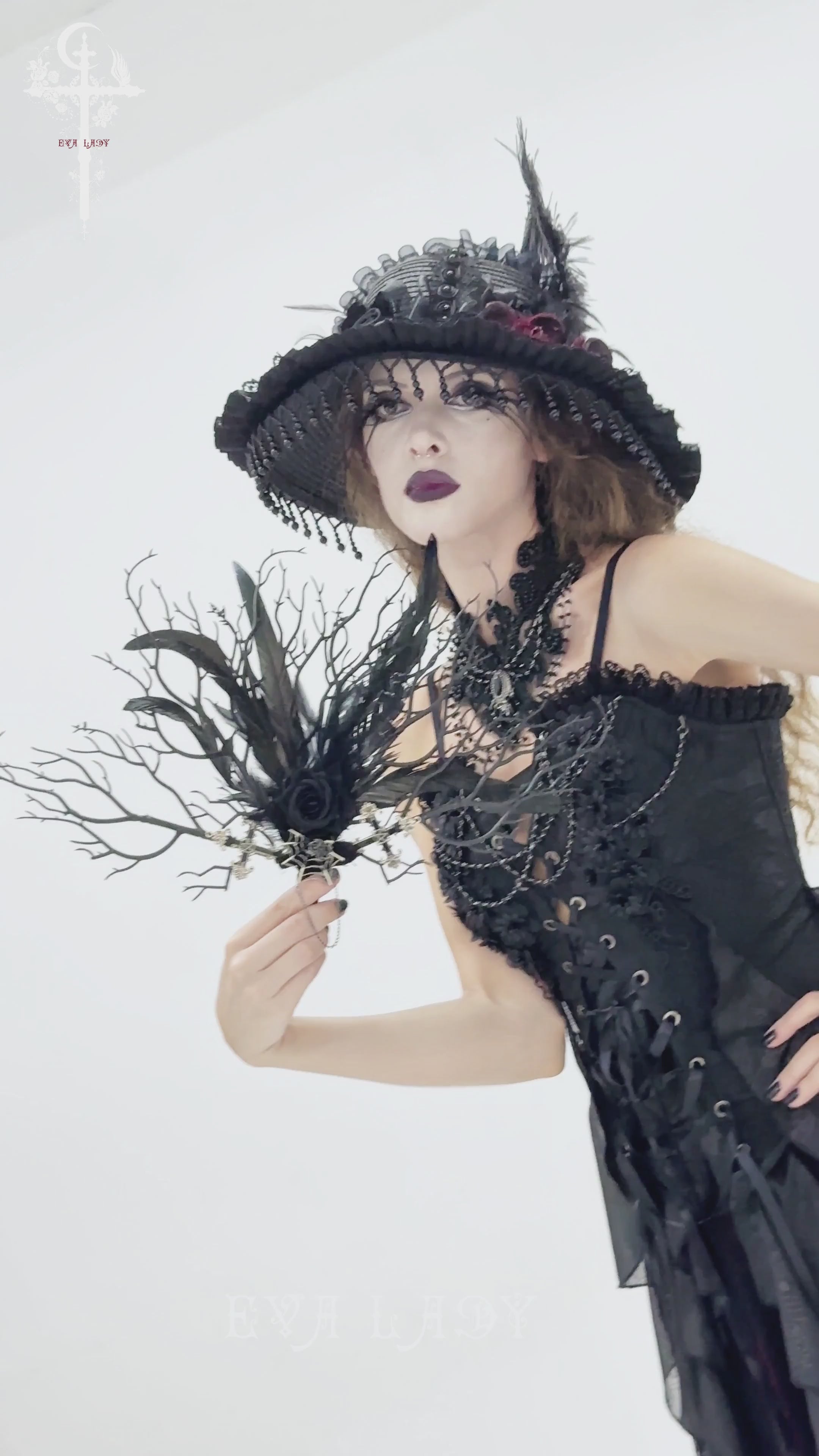 Model holding a Gothic Black Feather and Twig Hand Fan with Cross Charms, wearing a dramatic black corset and hat.
