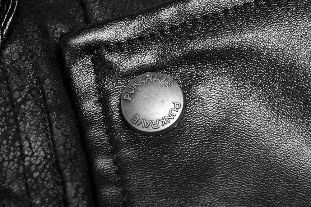 Close-up of a PUNK RAVE metal button on a black rubberized coat, showcasing edgy punk-inspired design.