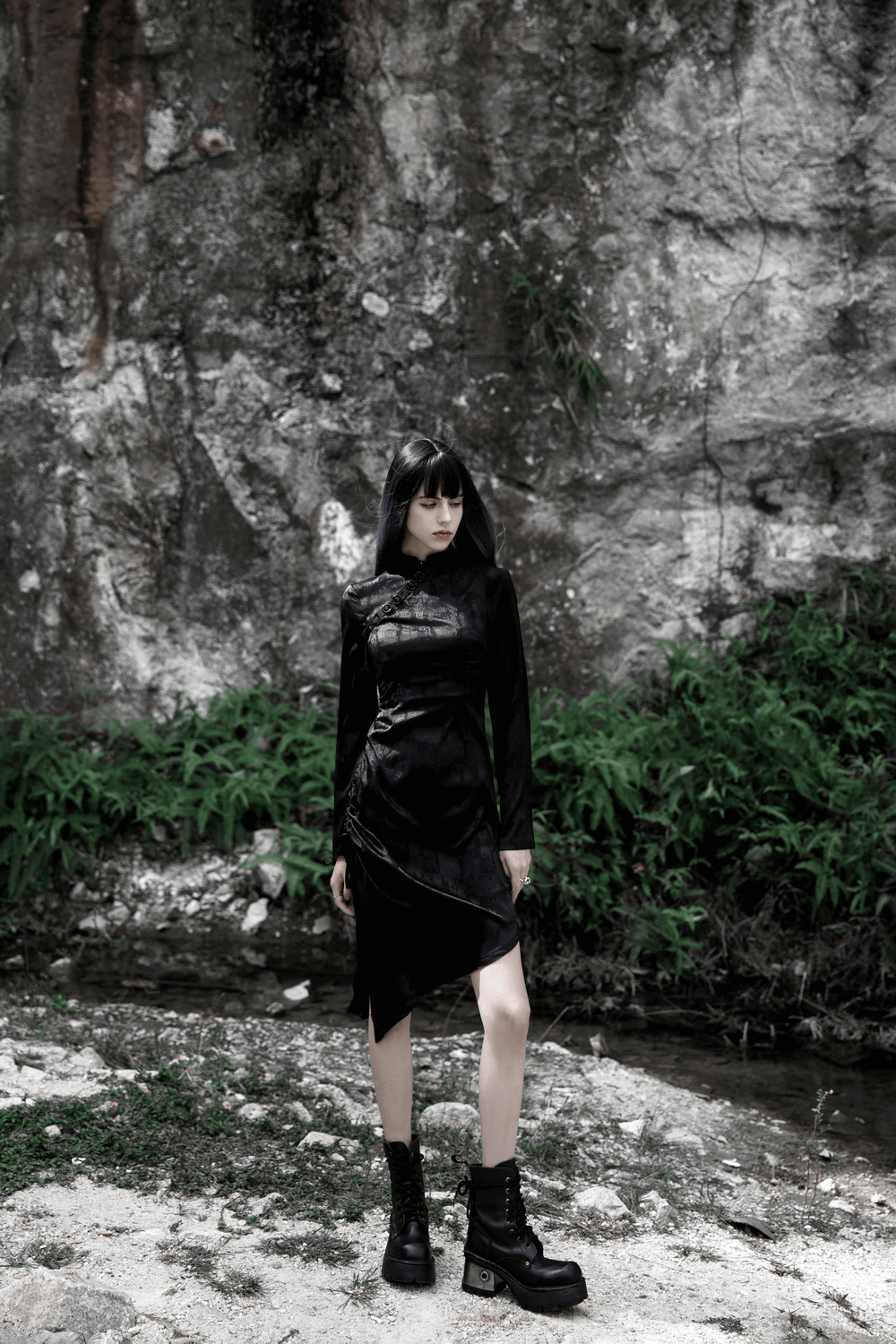 Gothic black cheongsam dress with pointed hem and long sleeves, model posing in nature with edgy boots.