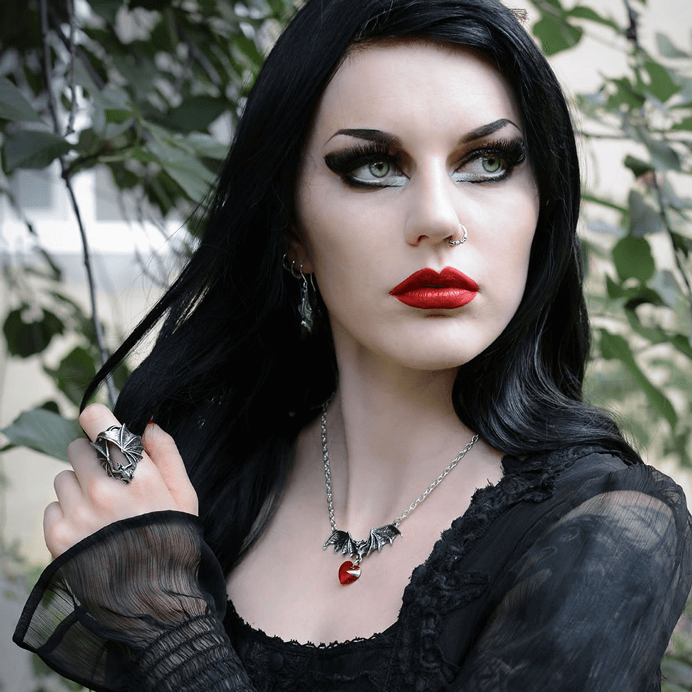 Model showcasing gothic style with Stealth Bat Ring and bat-themed necklace, set against a lush green background.