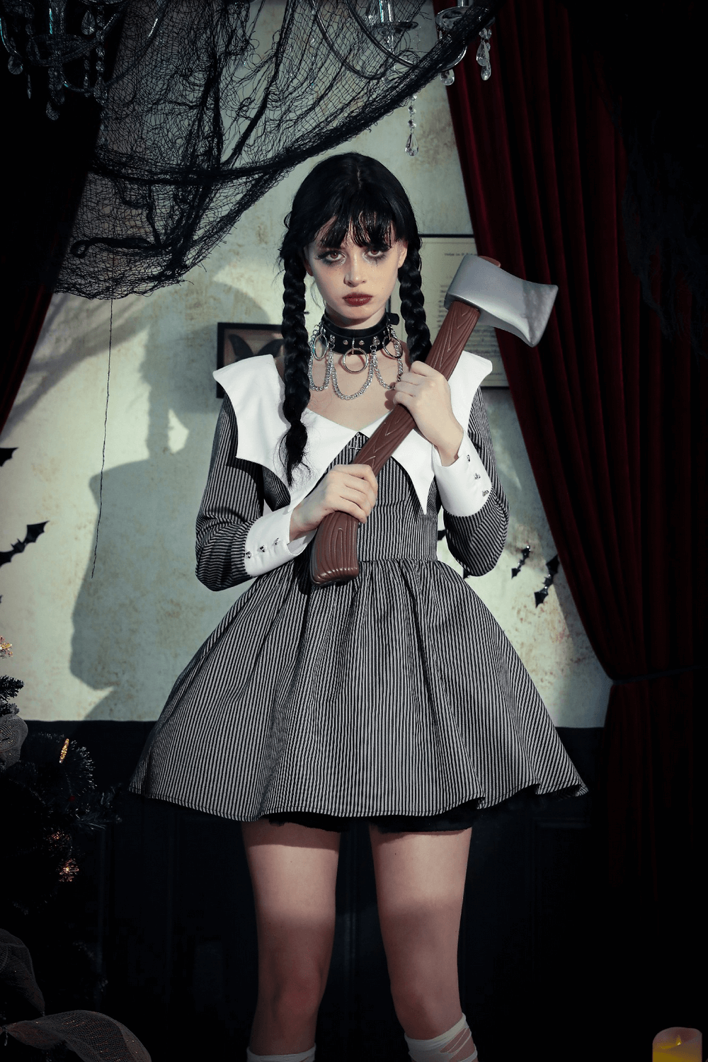 Model in striped dress with white collar, holding an axe, set in a spooky environment with dark curtains and decorations.