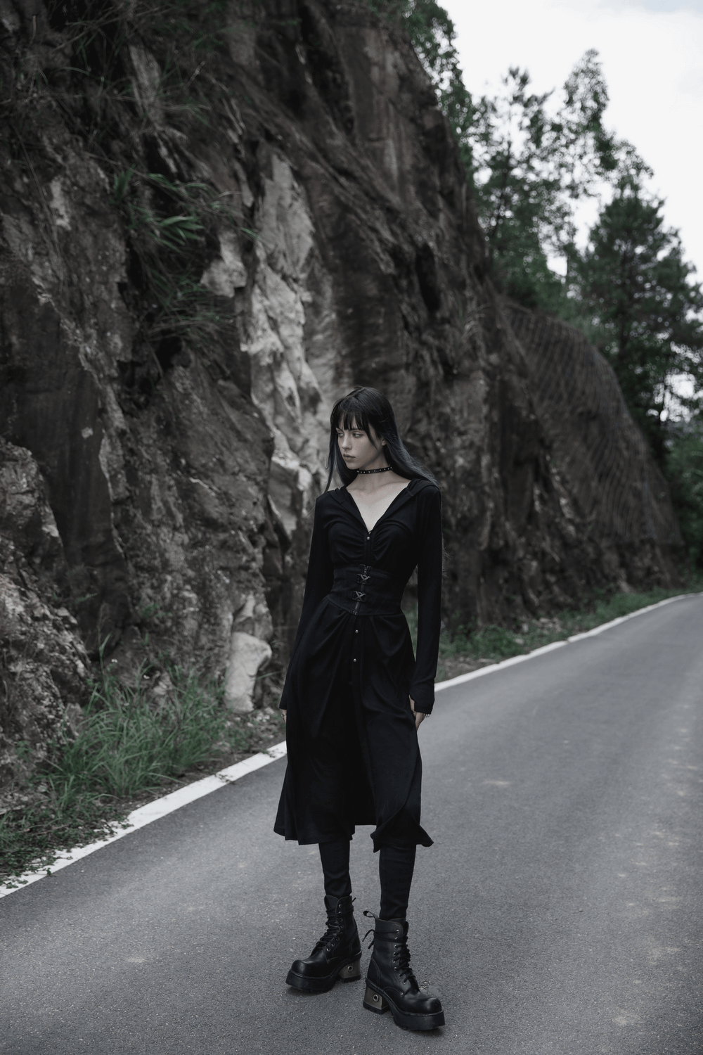 Model in sleek black outfit standing on a road surrounded by rocky terrain, showcasing modern edgy fashion.