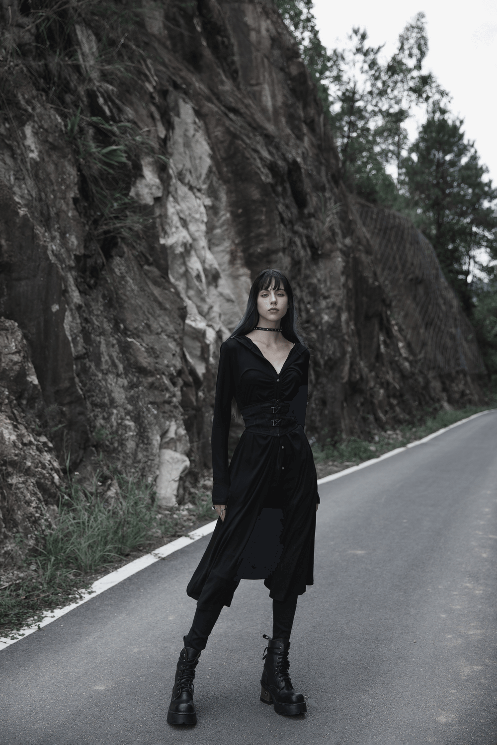 Model in black outfit stands on roadside with rocky backdrop, showcasing edgy fashion and striking style.