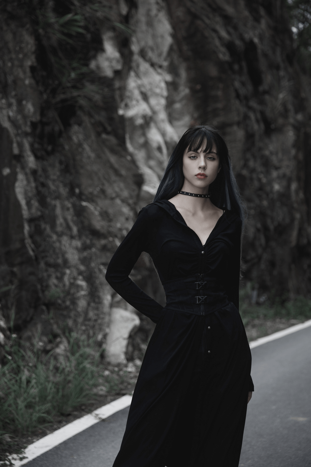 Model in black outfit with waist-cinching buckle detail, posing against a rocky backdrop.