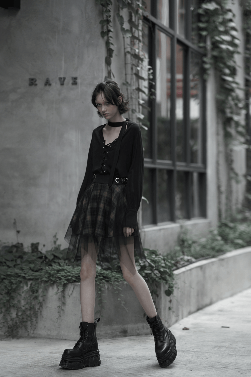 Edgy fashion model in black mesh and plaid asymmetric skirt, showcasing punk style with layered detailing and combat boots.