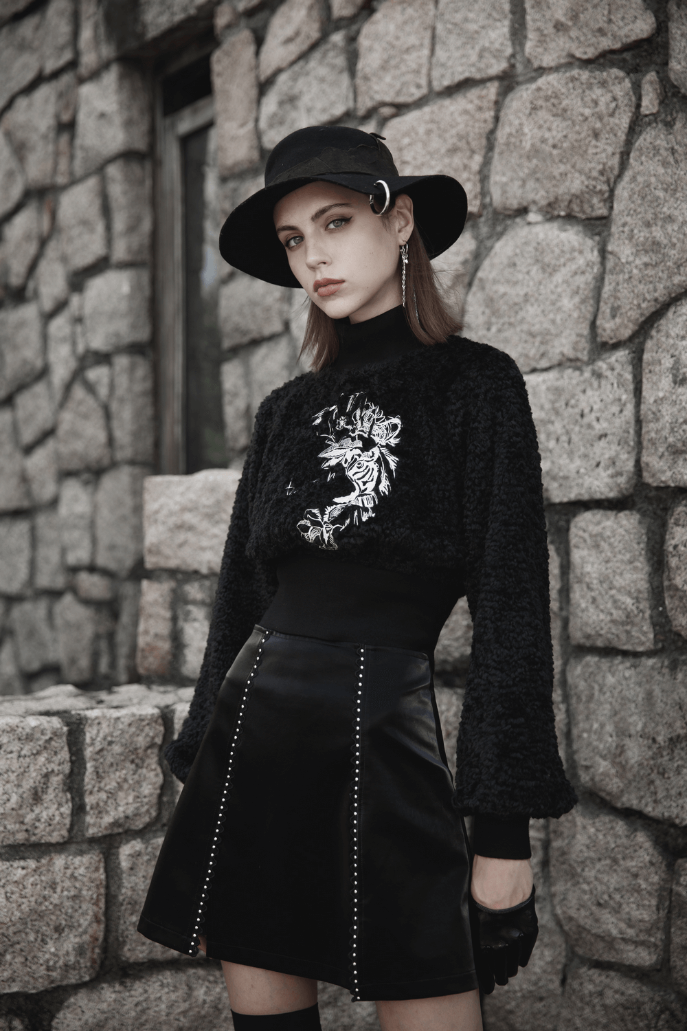 Model in a stylish black Gothic mini skirt with lacing, paired with an embroidered sweater and hat, against a stone wall.