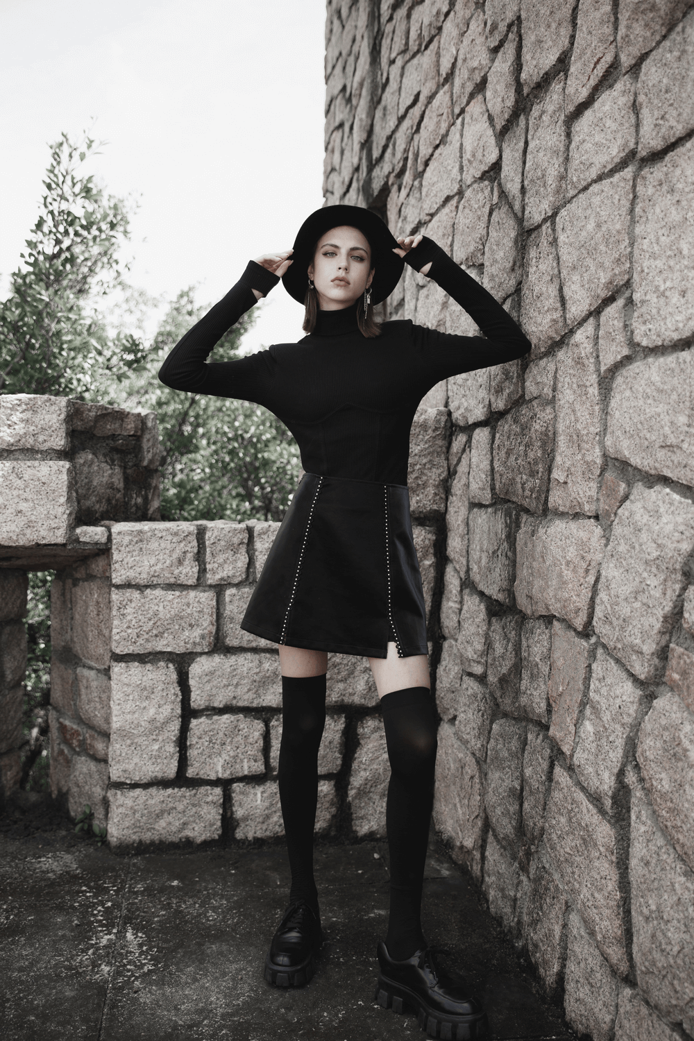 Stylish Gothic mini skirt with lacing and rivet details, paired with a black long-sleeve top and hat against a stone wall.