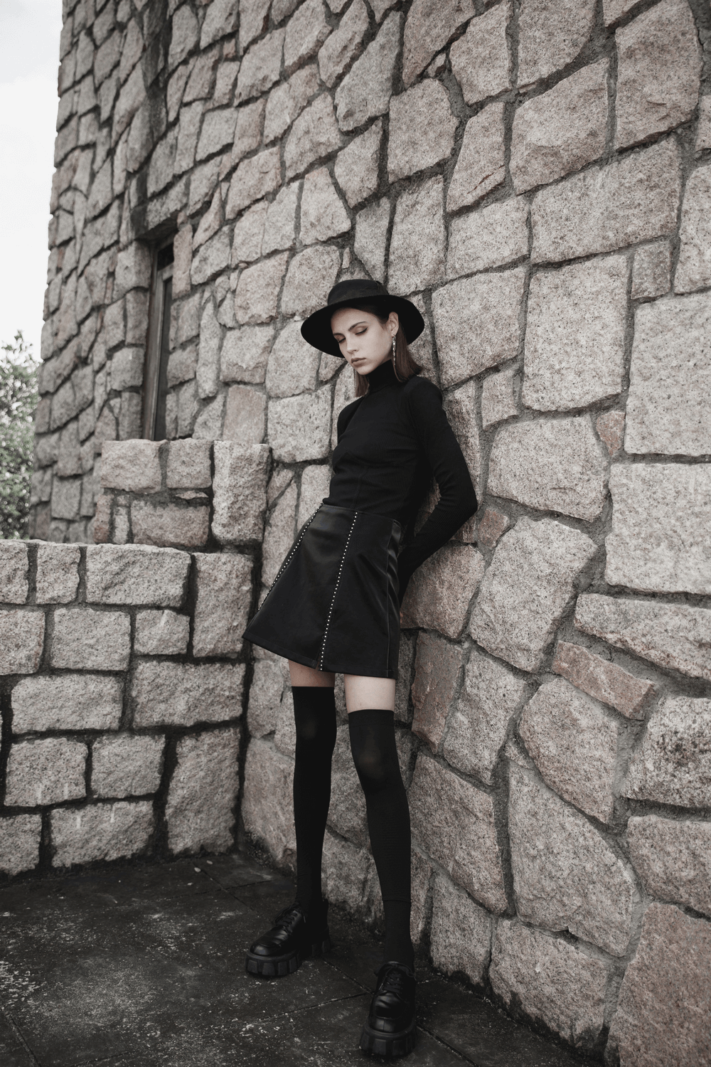 Gothic mini skirt with lacing, stylishly paired with a black top and hat, against a stone wall backdrop.