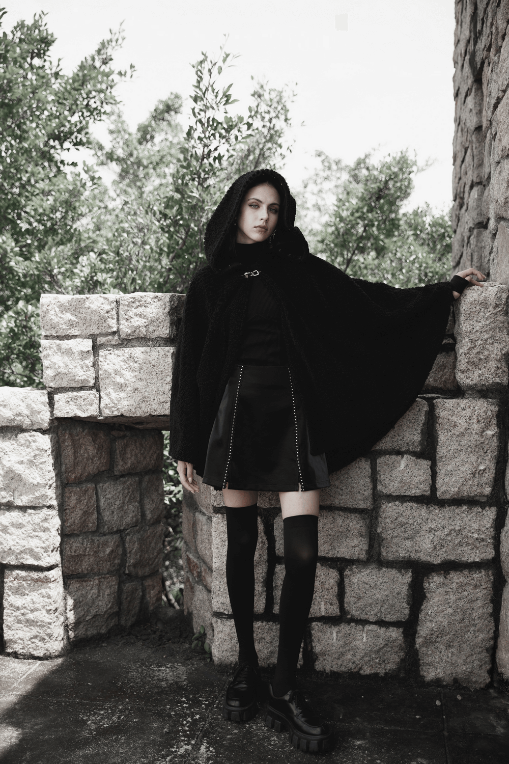 Model wearing a stylish black Gothic mini skirt and hooded cape, striking a confident pose outdoors.