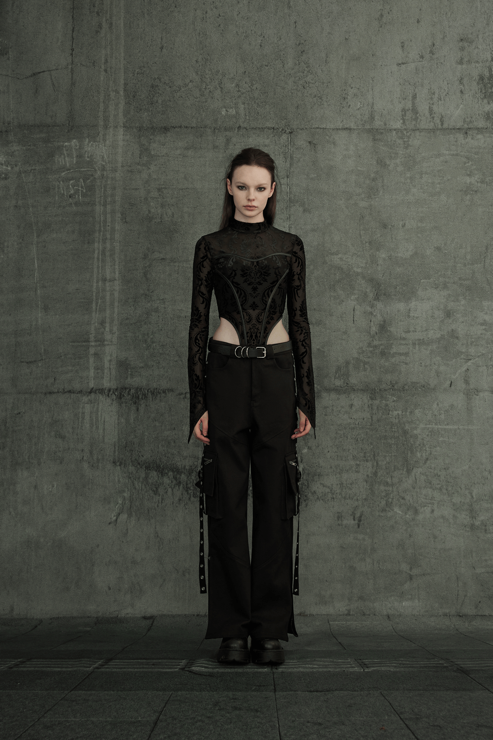 Model wearing a black sheer bodysuit and cargo pants against a concrete wall, showcasing modern streetwear style.
