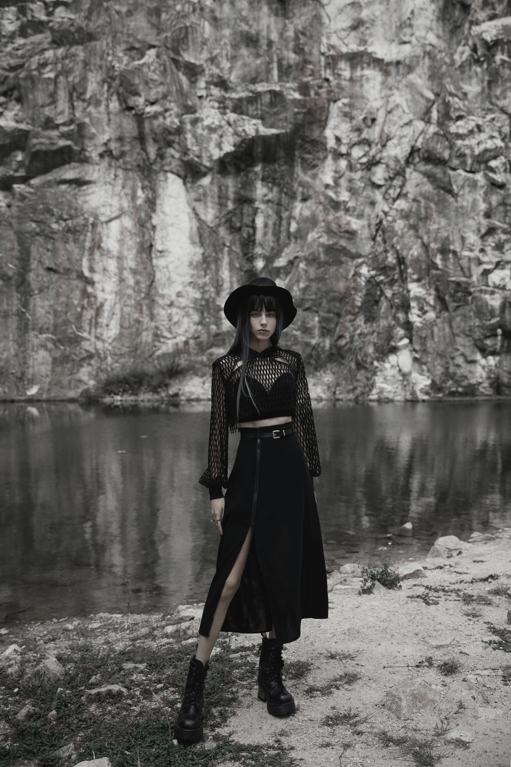 Model in a chic black A-line midi skirt with slit and hat, posing by a serene rocky lake background.
