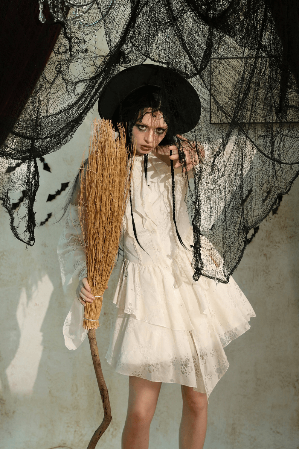 Model in a whimsical white Victorian lace dress holding a broomstick under dark, dramatic netting backdrop.