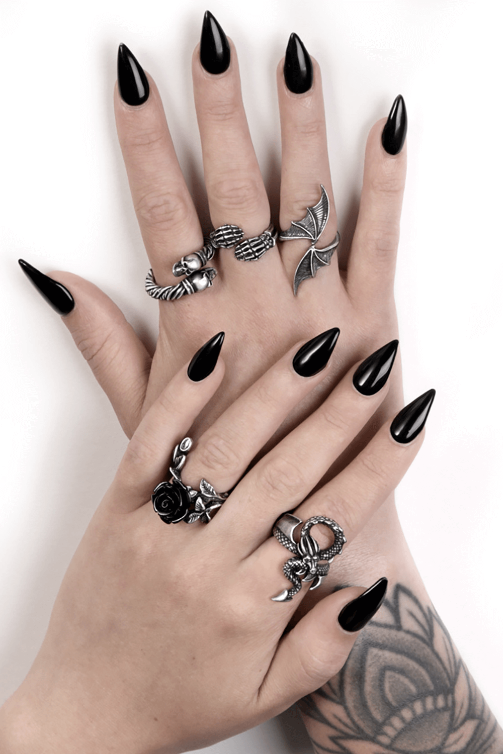Stylish hands adorned with silver rings, featuring intricate designs like a dragon, skulls, and roses, on glossy black nails.