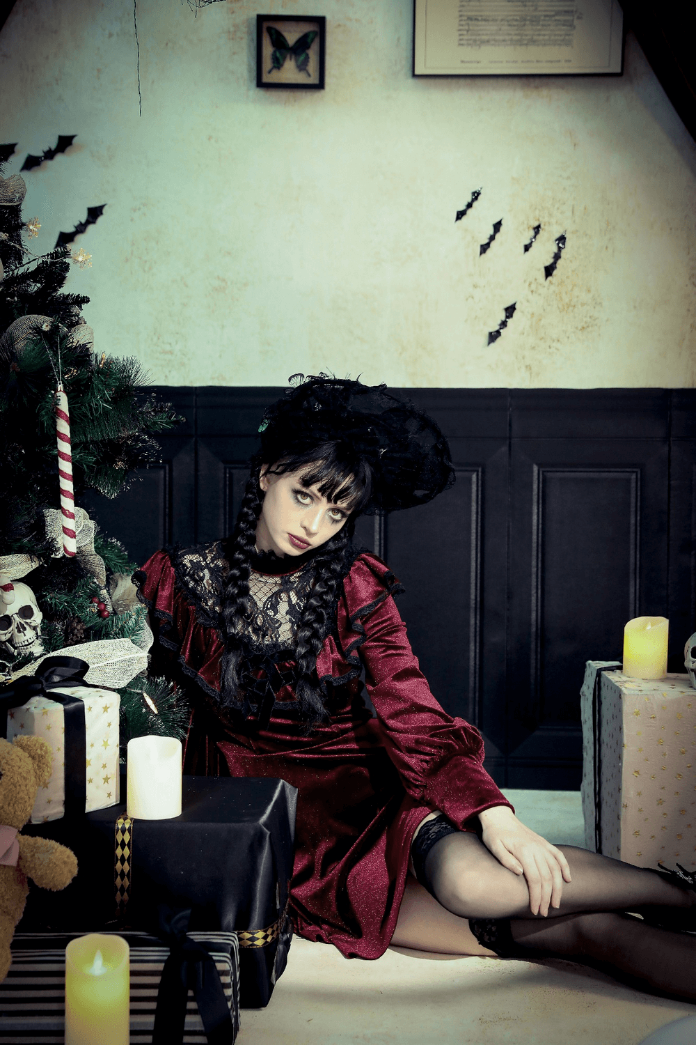 Model in a burgundy velvet gothic mini dress with black lace trim sits by a decorated Christmas tree.