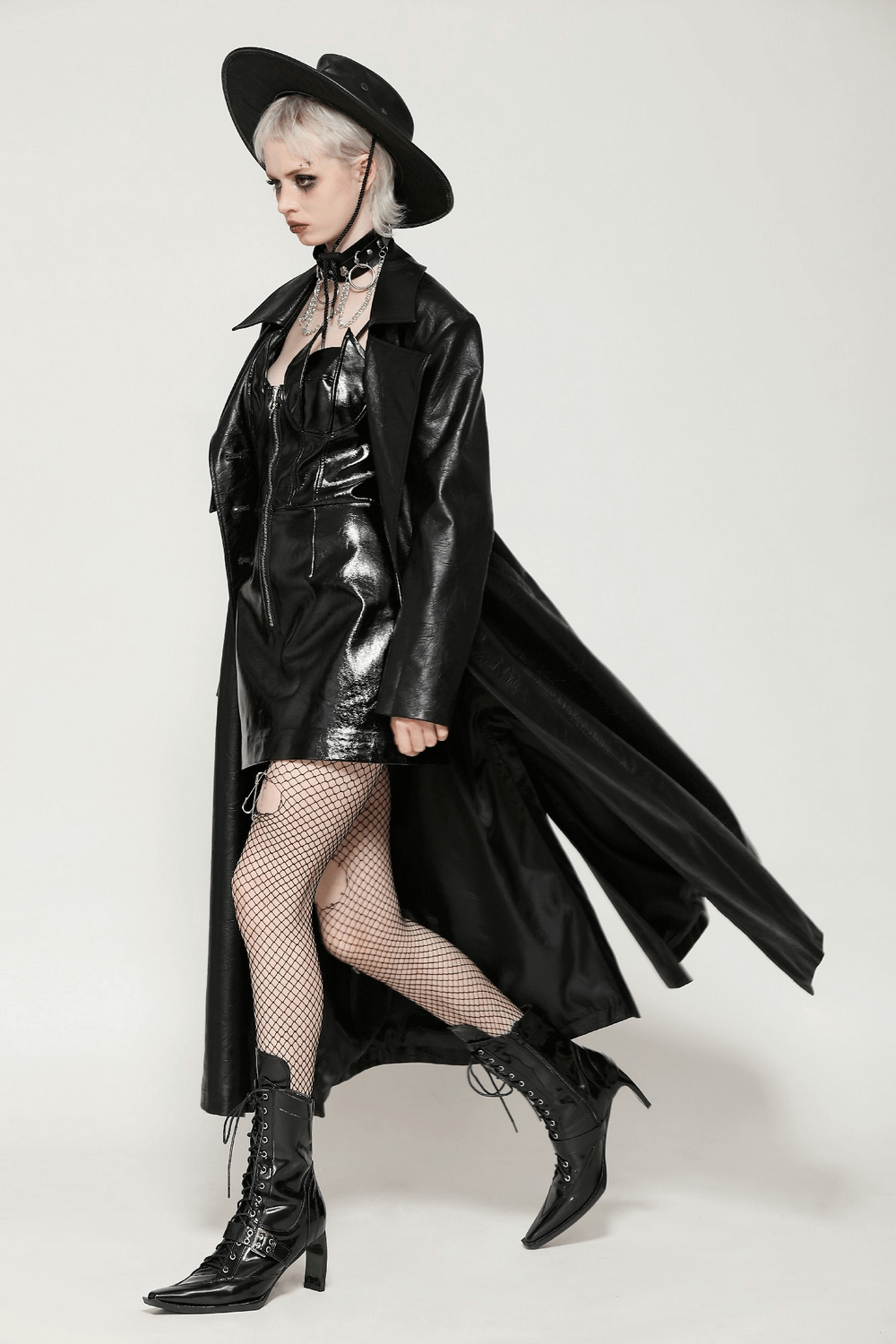 Woman wearing a long black faux leather trench coat with a belted waist and a hat, showcasing edgy fashion style.