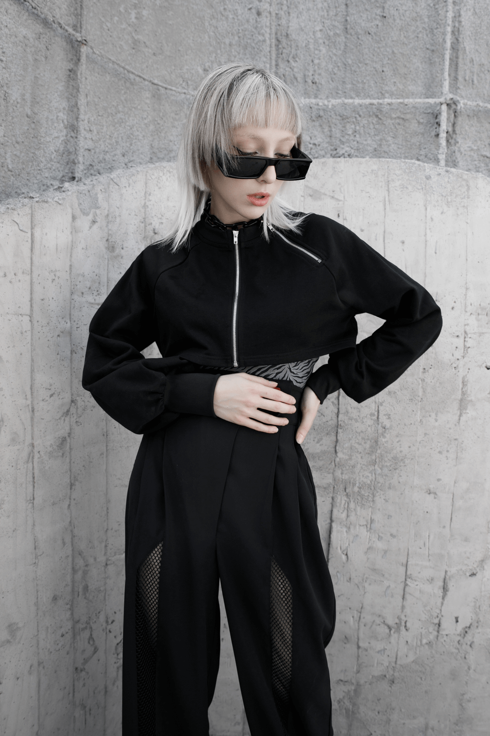 Edgy punk style black zip-up crop jacket with chain detail, styled with trendy sunglasses and high-waisted pants.