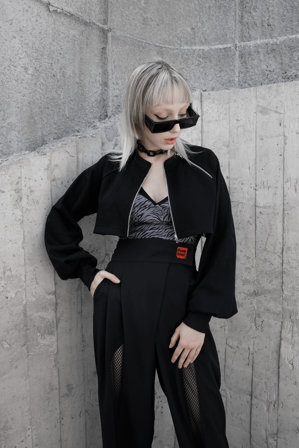 Model wearing a black zip-up crop jacket with long sleeves and chain detail, showcasing an edgy punk style.