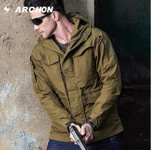 Men's military jacket in olive green with hood, worn by a man holding a firearm against a rugged backdrop.