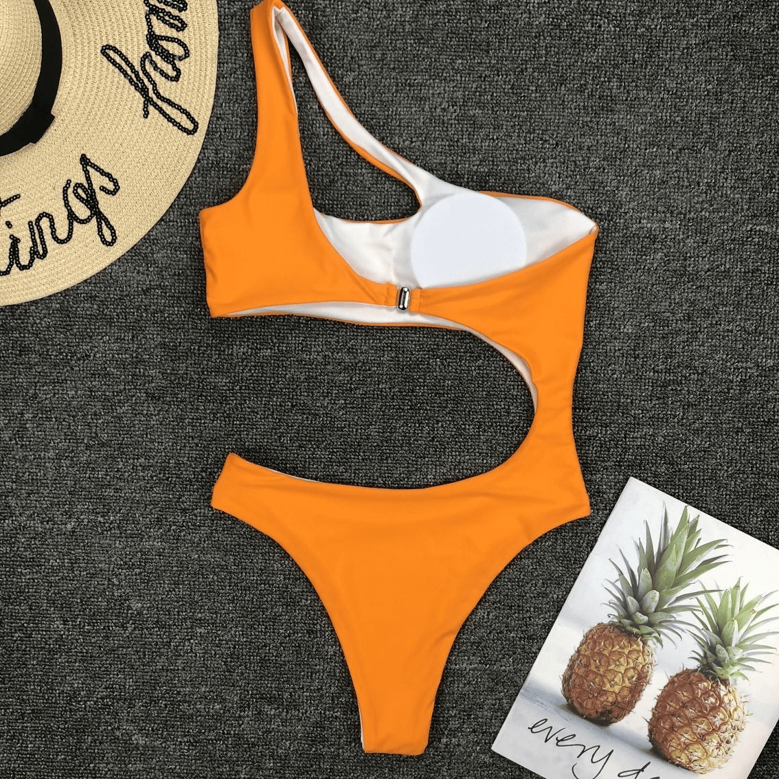 Stylish asymmetrical women’s orange one-piece swimsuit on a textured surface with beach accessories.
