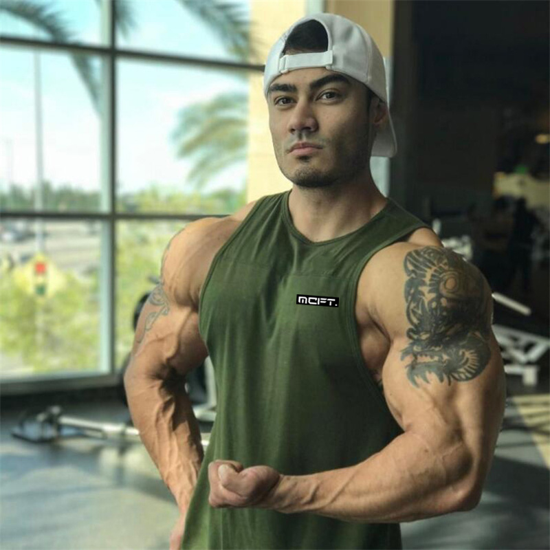 Muscular man wearing a green bodybuilding tank top posing in a gym with a bright window backdrop.