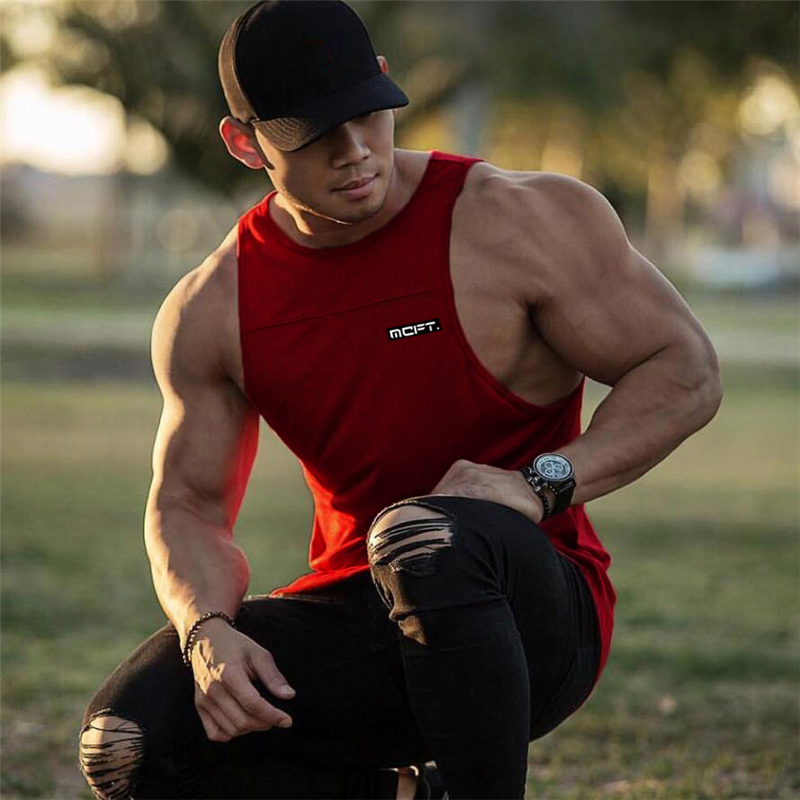 Muscular man wearing a red bodybuilding tank top, showcasing fitness style and casual look in a park setting.
