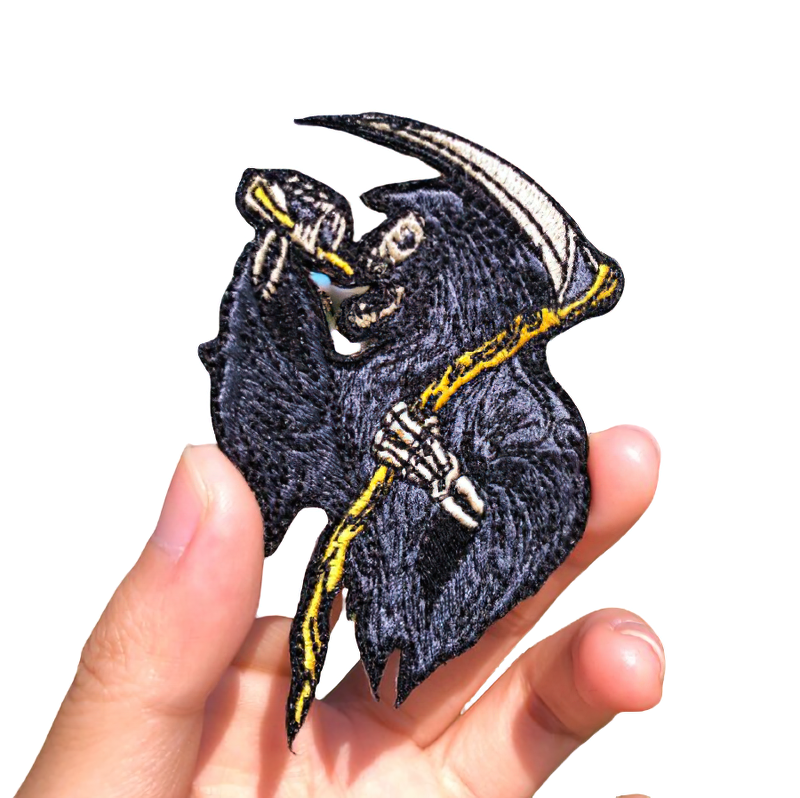 Hand holding a gothic patch of death with a scythe and drink, stylish accessory for jackets and bags.