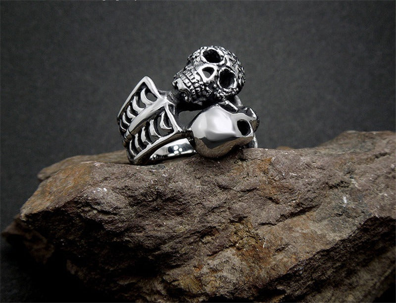 Double skull ring in vintage stainless steel, perfect for rocker and biker style, displayed on a textured rock background.
