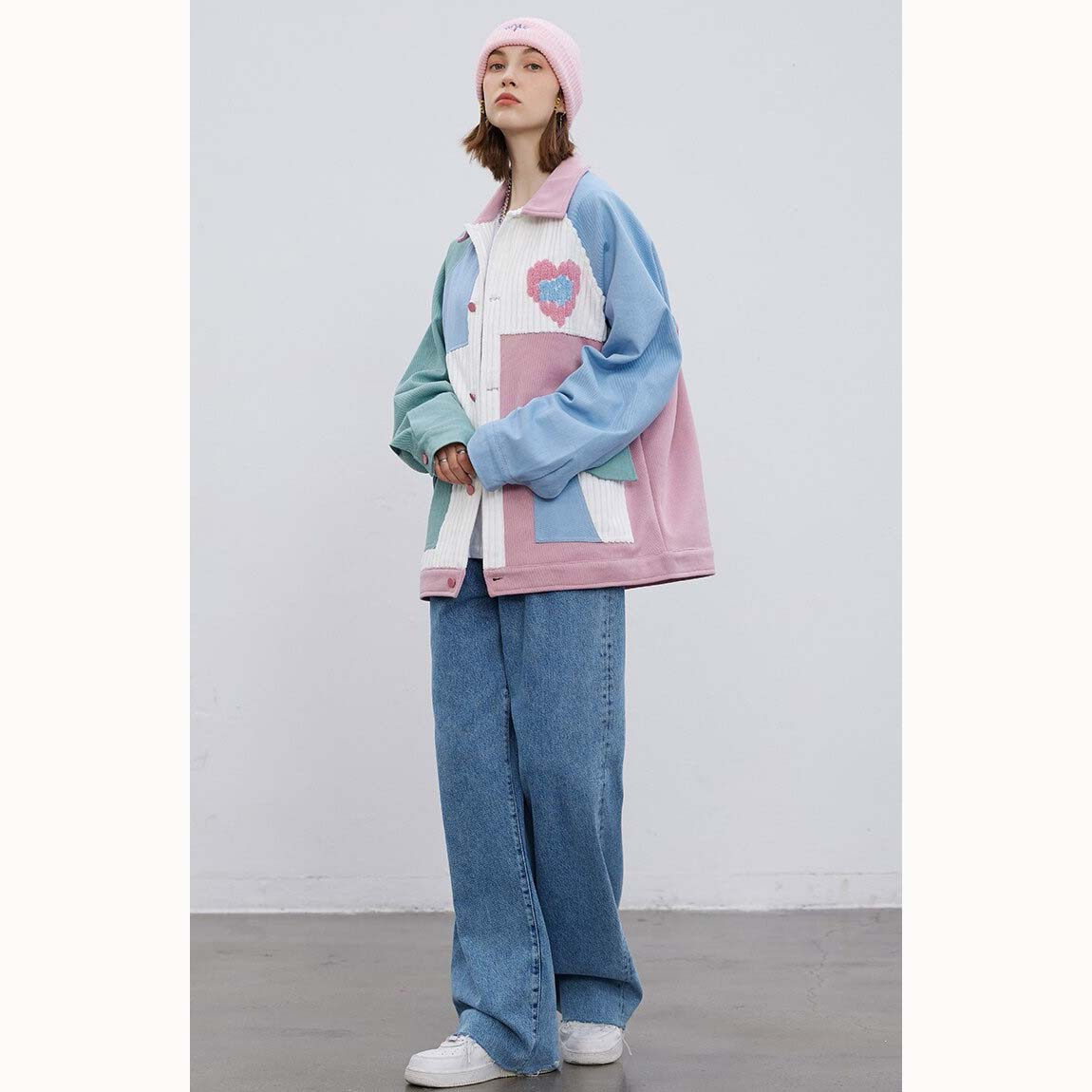 Stylish woman wearing oversized colorful patchwork jacket with heart details, paired with wide-leg jeans and pink beanie.