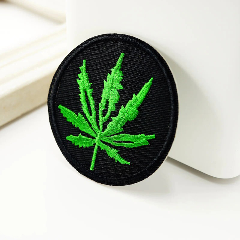Circular black patch featuring an embroidered green ganja leaf design, perfect for trendy clothing or accessories.