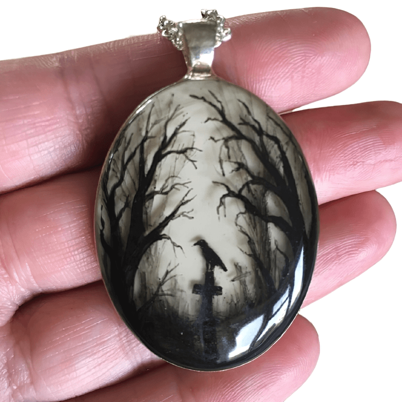 Hand holding gloomy wood graveyard pendant featuring a crow and bare trees in gothic style.