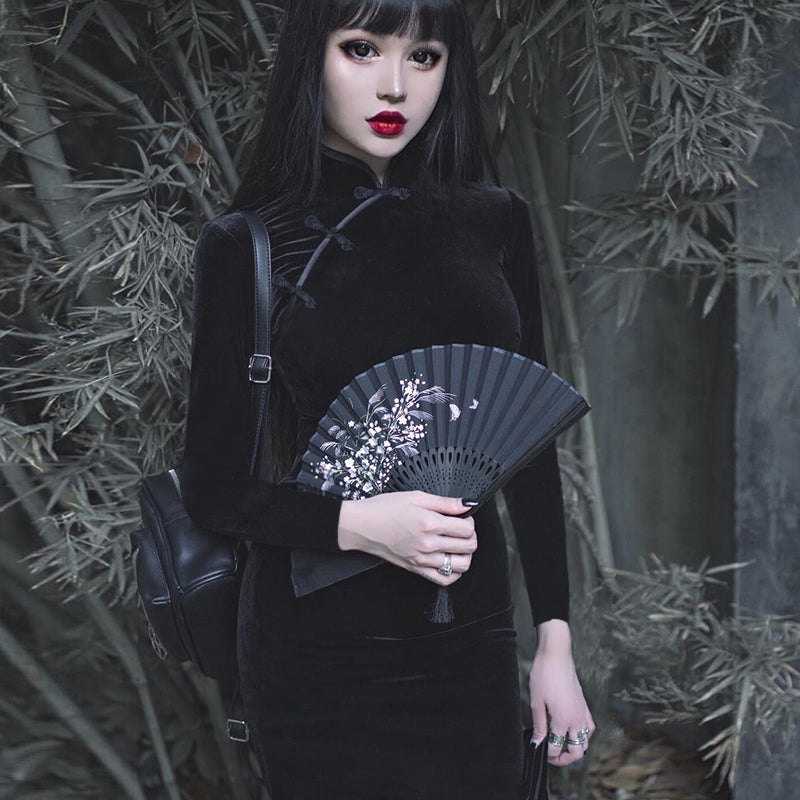 Elegant woman in a gothic velvet dress holding a black fan amidst bamboo, showcasing alternative fashion style.