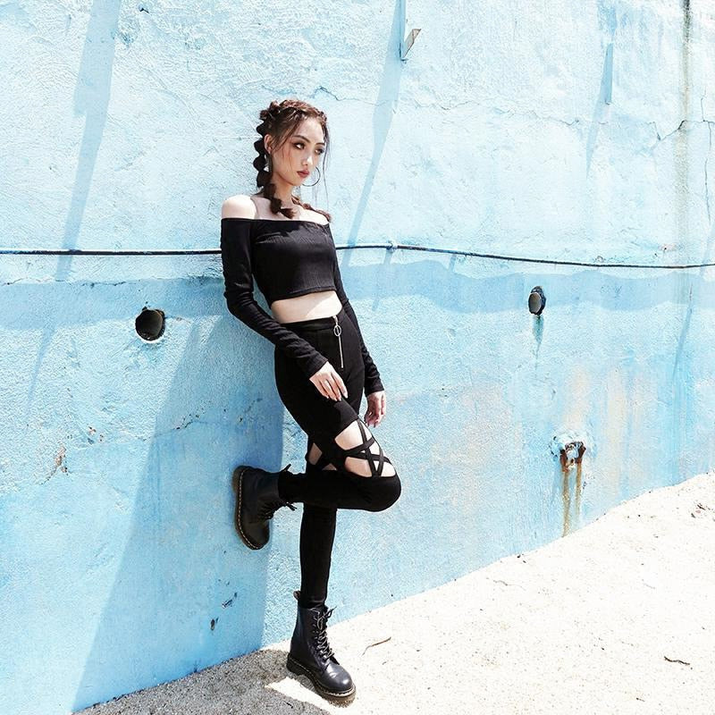 Gothic woman in black off-shoulder top and pencil pants with pentagram designs, posing against a blue wall.