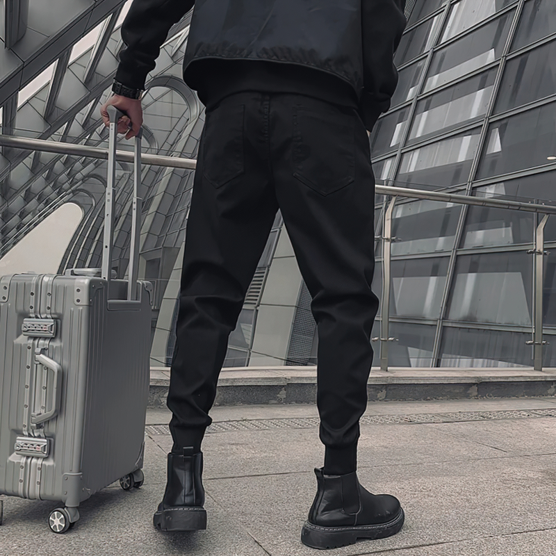 Stylish man in black drawstring cotton pants and boots, standing with luggage in a modern urban setting.
