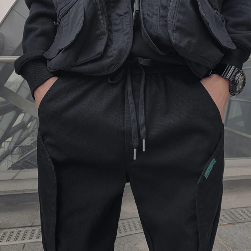 Men's stylish black cotton joggers with a drawstring, paired with a black vest in a modern urban setting.