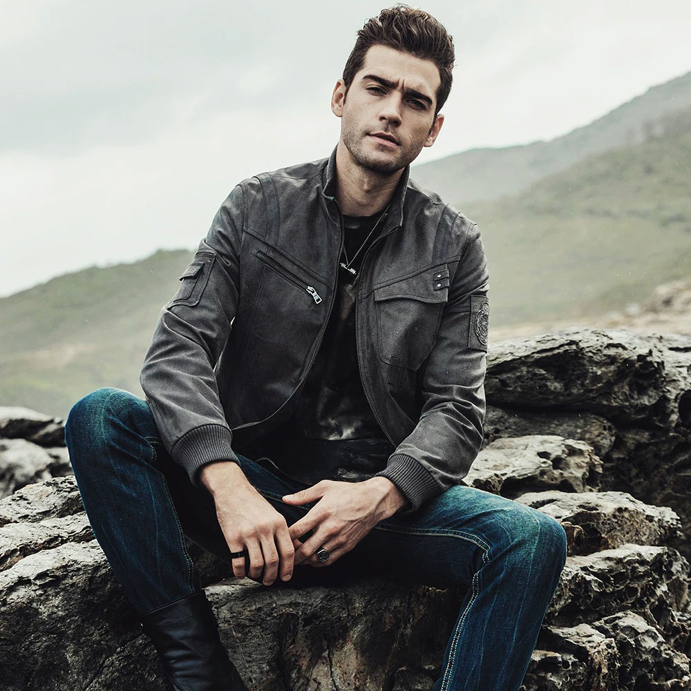 Stylish man in vintage real leather motorcycle jacket, seated on rocks with a rugged landscape in the background.