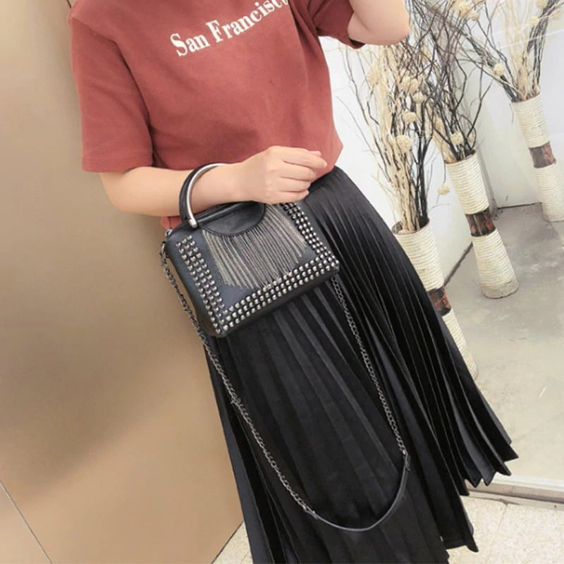 Stylish woman in a maroon top holding a rock-style rivet crossbody bag with chains and studs, paired with a pleated black skirt.