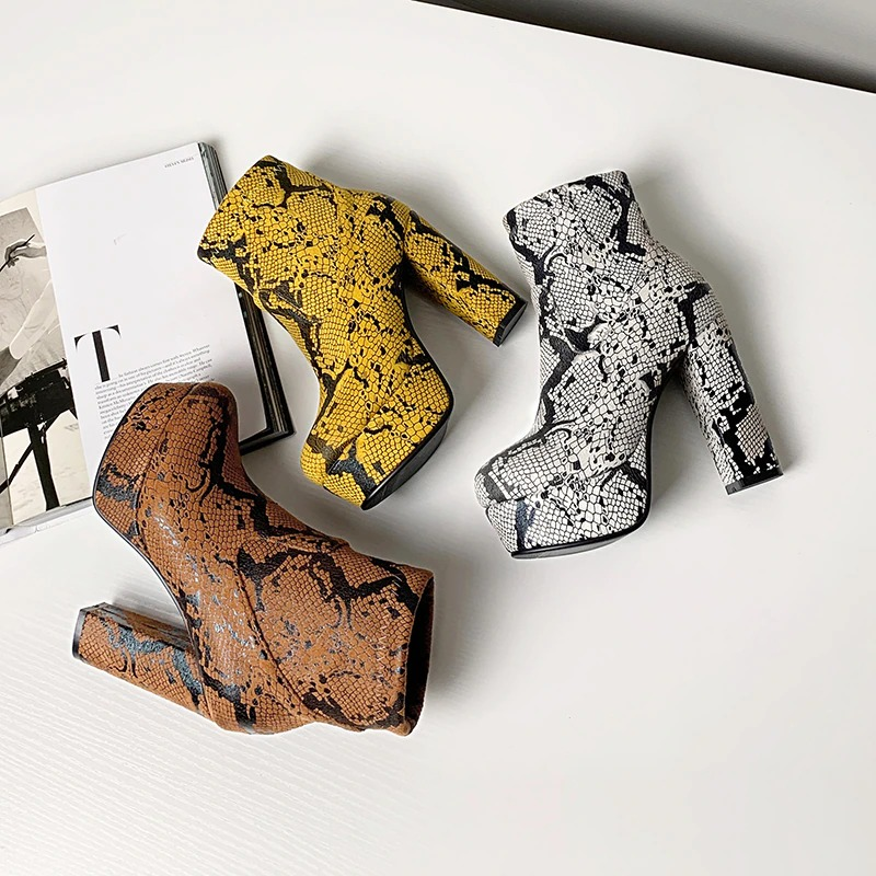 Trendy women's snake skin print ankle boots in yellow, gray, and brown on a stylish table backdrop.