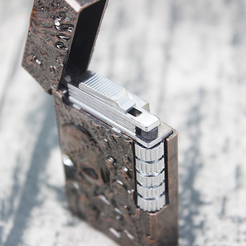 Skull gas lighter in a metallic finish, featuring embossed skull designs and a sleek opening mechanism.