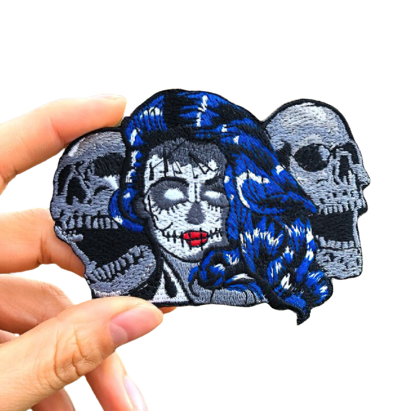 Hand holding a colorful skull patch featuring a woman with blue hair and two skulls, perfect for jackets and bags.