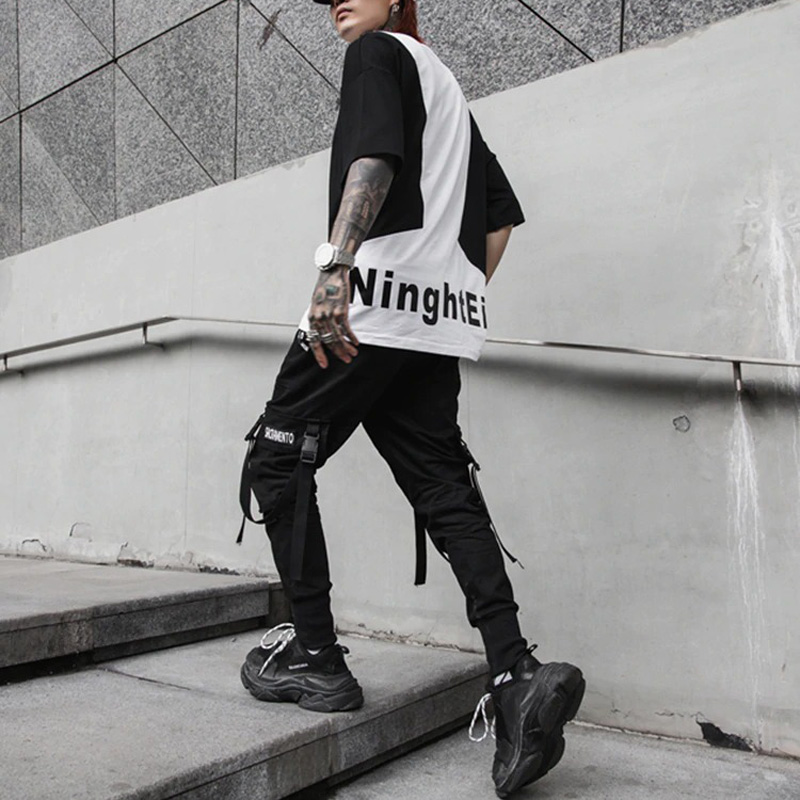 Stylish man wearing black spring fashion pants with ribbons and a graphic tee, walking confidently on urban steps.
