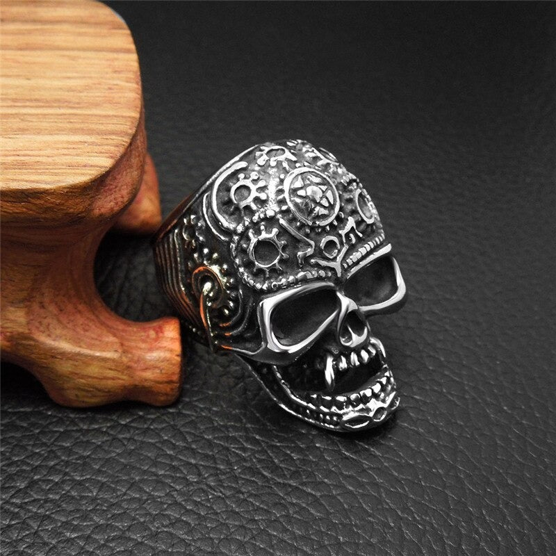 Stainless steel big skull ring with detailed gear design on a wooden display against a black background.