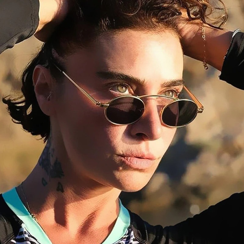 Stylish woman in small oval sunglasses with a steampunk flair against a natural background, displaying UV400 protection.