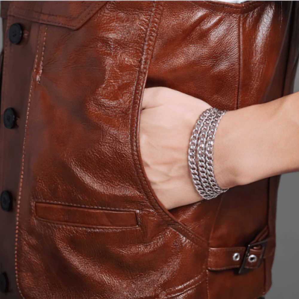 Close-up of a hand wearing a silver chain bracelet, tucked in the pocket of a vintage brown leather biker vest.
