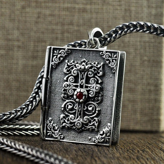 Vintage silver religious pendant shaped like a book, embellished with intricate details and a striking red gemstone.