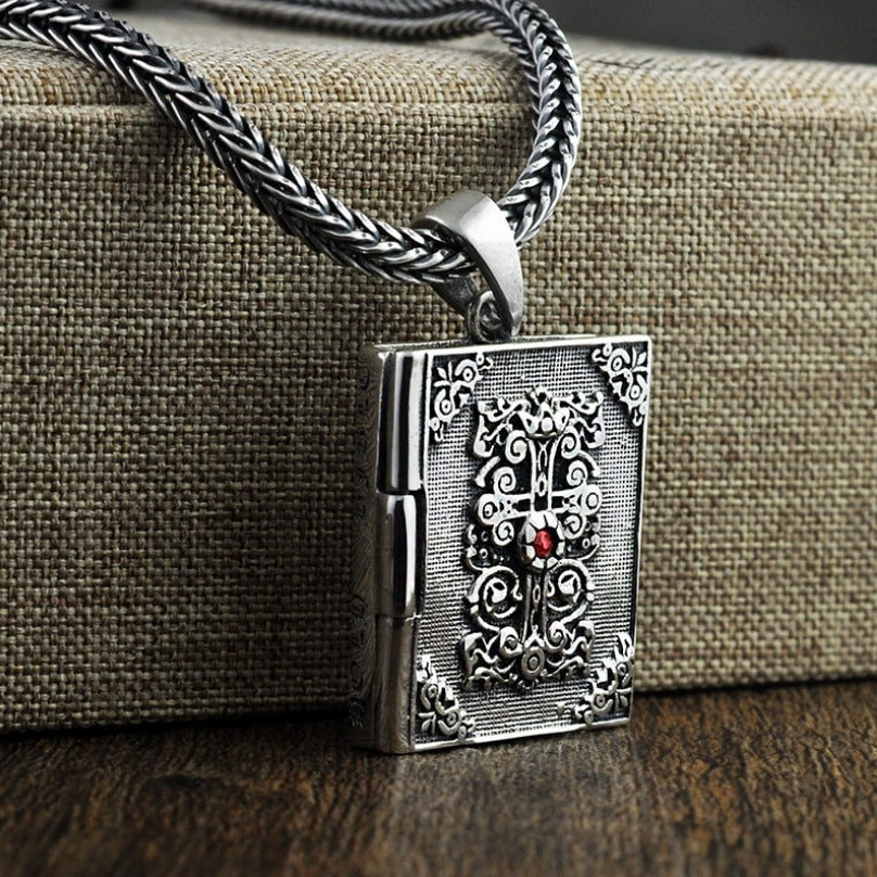 Vintage silver pendant shaped like a religious book, featuring intricate detailing and a red gemstone.